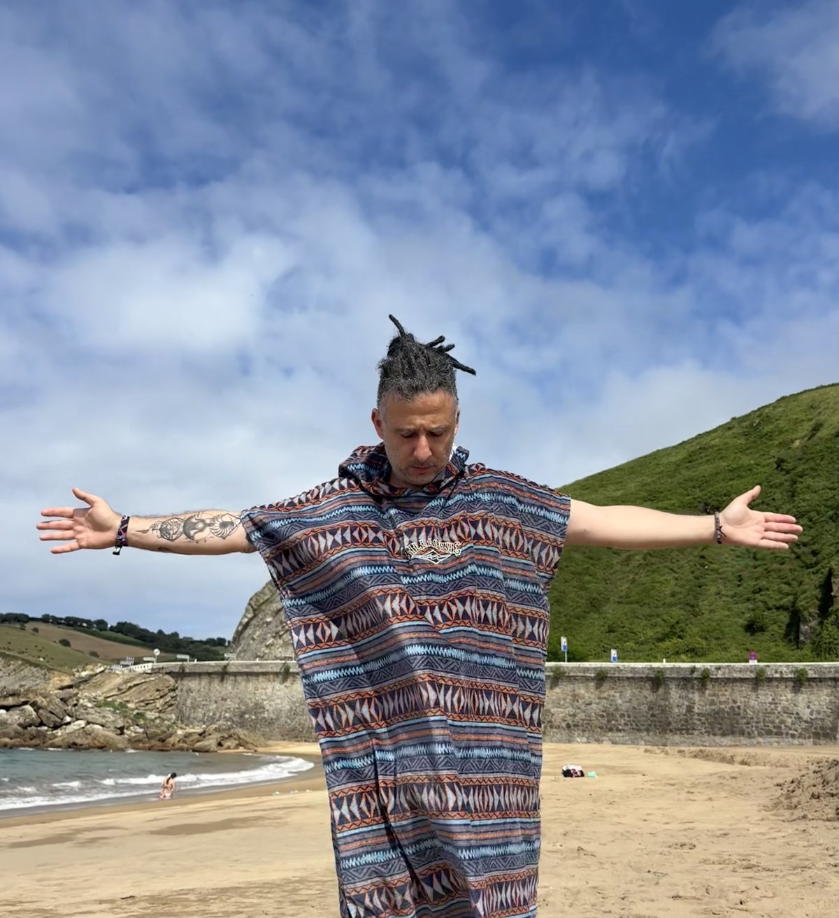 TomaTrip on a beach, arms wide, eyes closed, in a patterned poncho under a cloudy blue sky